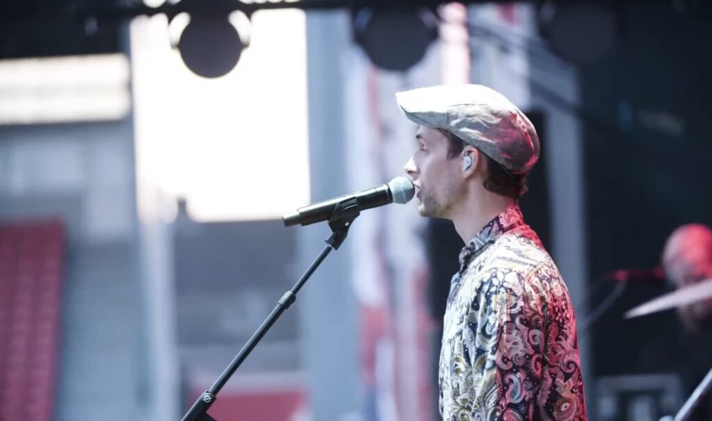  person is seen singing into a microphone on stage, wearing a patterned shirt on the stage